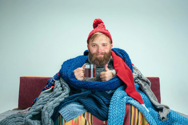 bearded sick man with flue sitting on sofa at home or studio with cup of tea covered with knitted warm clothes illness influenza concept relaxation at home healthcare concepts 155003 17523